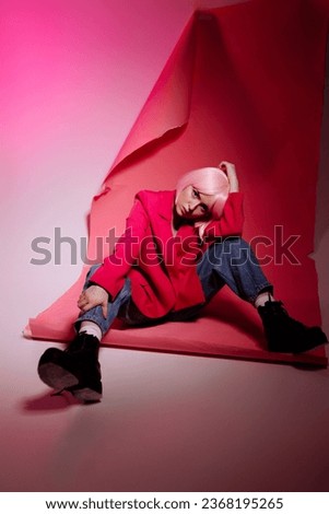 A stylish woman with pale pink bob hair wearing a pink jacket and jeans confidently poses on the floor against a pink background. Fashion advertising concept, individuality and confidence.