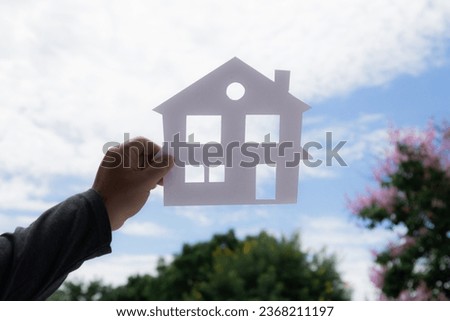 Woman Hand hold papercut of house picture against beautiful blue sky background, housing loan lifestyle concept, people dreaming or Stability in life, wanting to have a house, wealthy life planning