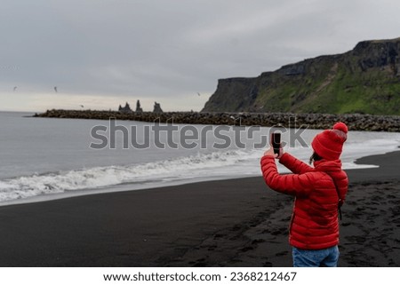 The girl takes pictures of the ocean on the phone. The girl is standing on a black beach. Iceland.