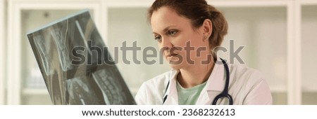 Doctor with stethoscope looks and analyzes x-ray of spine of patient sitting at table in office. Woman examines radiograph picture in clinic