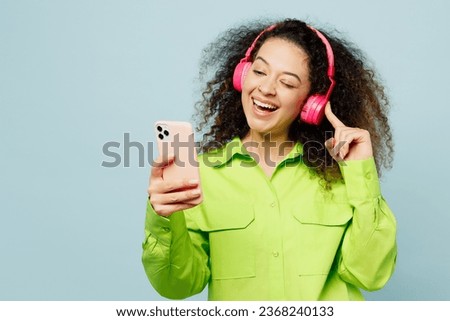 Young cool latin woman she wear green shirt casual clothes listen to music in headphones use mobile cell phone isolated on plain pastel light blue cyan background studio portrait. Lifestyle concept