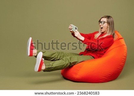 Full body young woman wear red shirt casual clothes glasses sit in bag chair use play racing app on mobile cell phone hold gadget smartphone for pc video game isolated on plain pastel green background