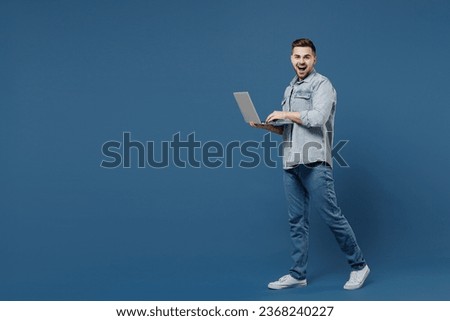 Full size length body side view profile laughing beautiful happy young brunet man 20s wears denim jacket hold use work on laptop pc computer go walk isolated on dark blue background studio portrait.