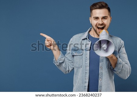 Fascinating smiling young brunet man 20s wears denim jacket hold scream in megaphone announces discounts sale Hurry up pointing aside away look camera isolated on dark blue background studio portrait.