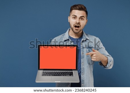 Shocked surprised stupefied young brunet man 20s wears denim jacket hold use work on laptop pc computer with blank screen workspace area point on it isolated on dark blue background studio portrait.