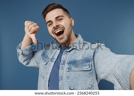 Close up excited young brunet man 20s wears denim jacket doing selfie shot on mobile phone showing thumb down dislike gesture isolated on dark blue background studio portrait. People emotions concept