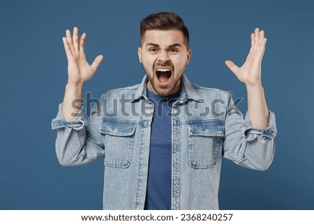 Distempered unnerved concerned aggrieved young brunet man 20s wears denim jacket spreading hands screaming shouting isolated on dark blue background studio portrait. People emotions lifestyle concept Royalty-Free Stock Photo #2368240257