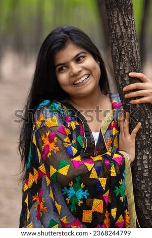 A pretty Indian woman in colorful traditional dress smiling and looking away holding a tree Royalty-Free Stock Photo #2368244799