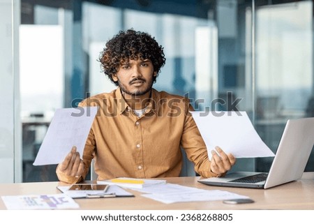 Portrait of young dissatisfied and disappointed financier, businessman behind paperwork looking at camera, negative checking documents and financial reports inside office, using laptop at work. Royalty-Free Stock Photo #2368268805