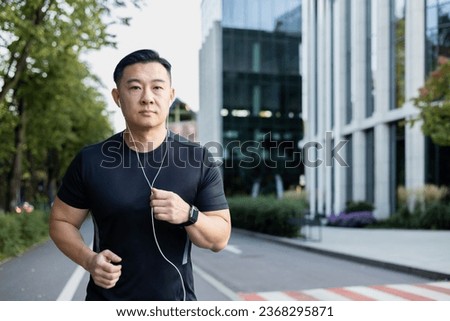 Portrait of a young Asian sportsman man running outside wearing headphones, listening to music, seriously looking at the camera.