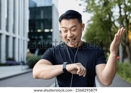 Close-up photo. Young asian male sportsman standing on city street after running and looking at smart watch on hand, happy with result.