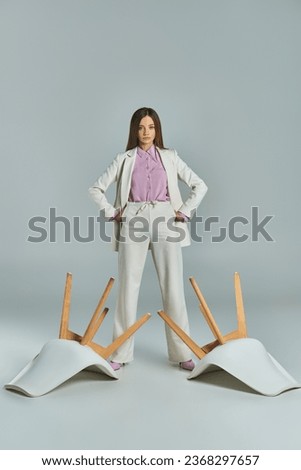 confident businesswoman in white suit standing with hands on hips near overturned armchairs on grey