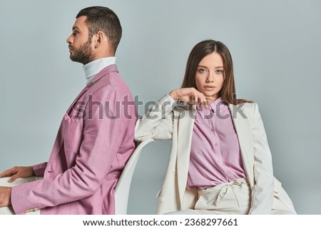 woman in elegant suit sitting in armchair and looking at camera near man in lilac blazer on grey Royalty-Free Stock Photo #2368297661