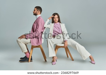 young woman in white suit sitting in armchair and looking at camera near man in lilac blazer on grey Royalty-Free Stock Photo #2368297669