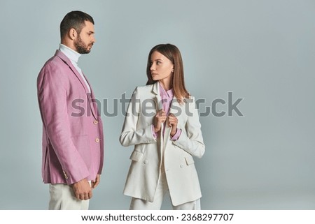 elegant woman in white suit looking at confident man in lilac blazer on grey, modern business couple Royalty-Free Stock Photo #2368297707