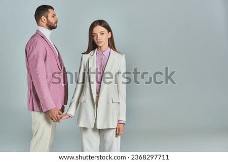young woman in white suit holding hands with man in lilac blazer and looking at camera on grey Royalty-Free Stock Photo #2368297711