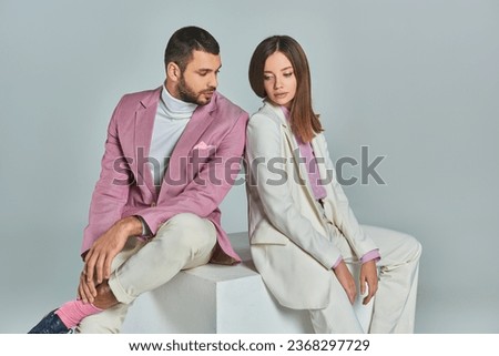 young and fashionable couple in elegant suits sitting on white cube on grey backdrop in studio Royalty-Free Stock Photo #2368297729