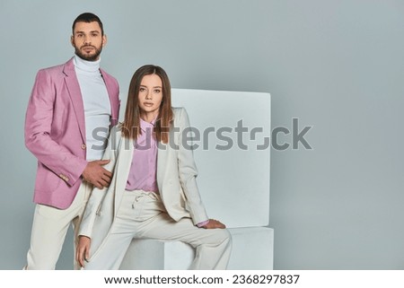 woman in white suit and man in lilac blazer looking at camera near white cubes on grey, trendy wear Royalty-Free Stock Photo #2368297837