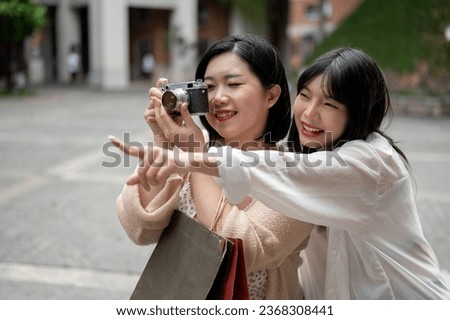 Two happy young Asian female friends are enjoying shopping and strolling around the city together on the weekend, using a film camera to take a picture of the city. Urban lifestyle, tourist, vacation
