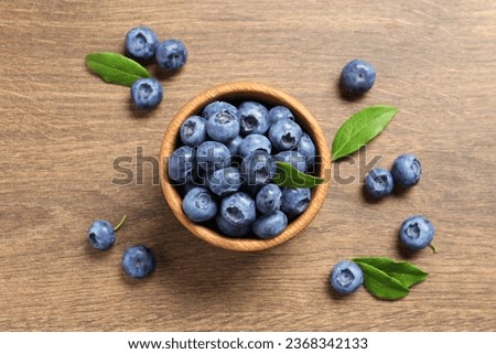 Bowl of tasty fresh blueberries with green leaves on wooden table, flat lay