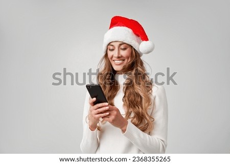 Woman christmas Santa Hat white sweater white studio background with smartphone in hand Beautiful caucasian female curly hair portrait. Happy person positive emotion Holiday concept Teeth smiling 