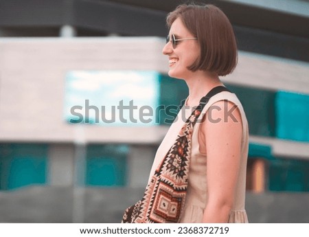 A young smiling happy girl, a successful woman in sunglasses on the background of a modern building walks through the city streets at sunset.