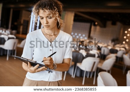 concentrated event organizer writing checklist on clipboard near festive tables in banquet hall Royalty-Free Stock Photo #2368385855