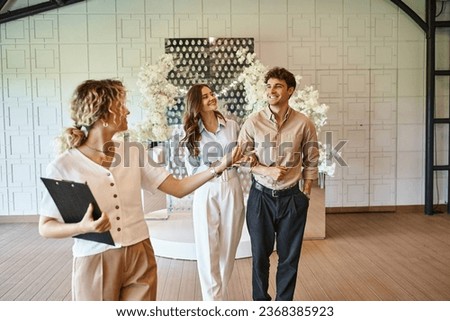 professional event manager with clipboard showing wedding venue with floral decor to happy couple Royalty-Free Stock Photo #2368385923