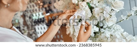 cropped view of decorator tying ribbon on white floral composition in festive event hall, banner Royalty-Free Stock Photo #2368385945