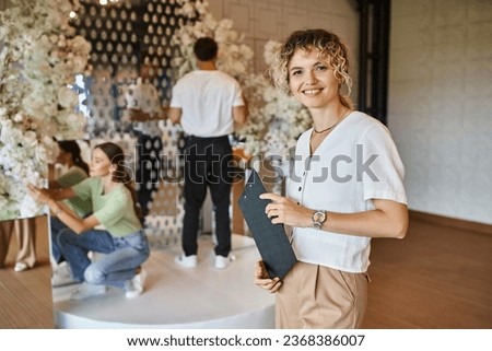 happy chief decorator looking at camera while team working with floral decor in event hall Royalty-Free Stock Photo #2368386007