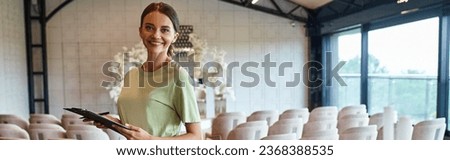 joyful event coordinator with clipboard smiling at camera in decorated banquet event hall, banner Royalty-Free Stock Photo #2368388535