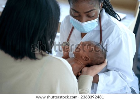 African pediatrician doctor wearing protective mask examines newborn baby in mother's arms Royalty-Free Stock Photo #2368392481
