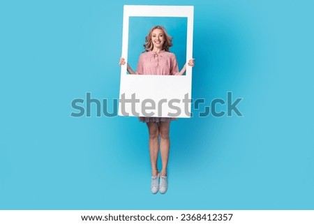 Full size photo of lovely positive girl dressed stylish pink clothes jumping holding frame in hands isolated on blue color background