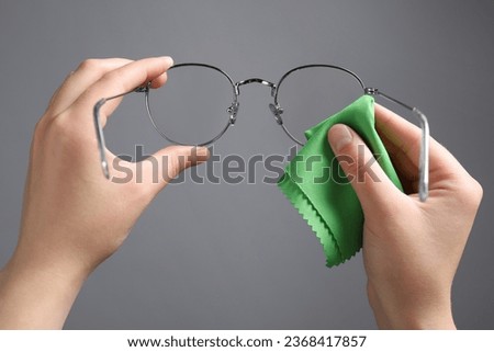 Woman wiping glasses with microfiber cloth on light grey background, closeup
