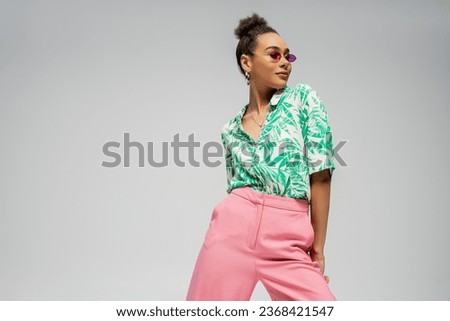 pleased african american woman in pink sunglasses and stylish attire posing on grey backdrop Royalty-Free Stock Photo #2368421547