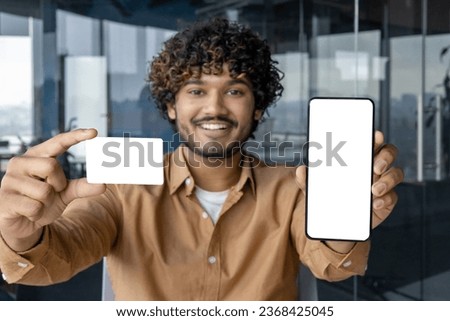 Portrait of young smiling Indian man sitting in office and holding mockup of mobile phone and credit card, showing white screen to camera.