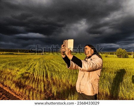 An adult girl in a field and with a stormy sky with clouds takes pictures of a rainbow and takes a selfie in the rain. A woman having fun outdoors on rural and rustic nature