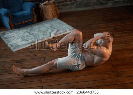 Endurance workout. Strong muscular man pumps abdominal muscles at home interior Caucasian athlete do morning workout indoor raises legs training abs muscle  Royalty-Free Stock Photo #2368443755