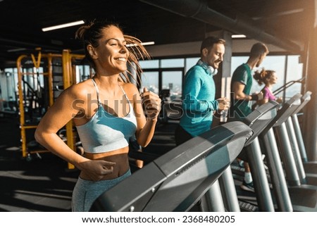 Happy athletic people jogging on treadmills in a health club. Beautiful people bathed in late afternoon sun running in a fancy gym. Selective focus of smiling slim fit woman exercising with friends.