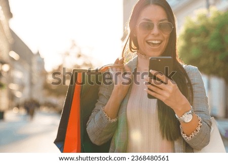 Attractive smiling woman with sunglasses checking bank account on her smart phone while shopping outdoors on a sunny afternoon.