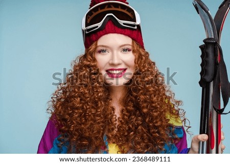 Smiling attractive curly woman with red lips wearing holding skis, wearing ski goggles, overalls, looking at camera isolated on blue background. Winter sports concept