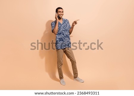 Full size photo of cool man wear print shirt indicating at sale empty space talking on smartphone isolated on beige color background