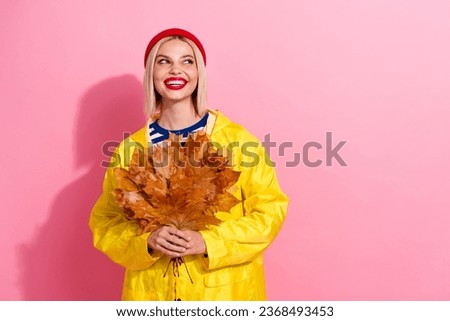 Photo of positive dreamy woman dressed rain coat gathering yellow leaves bouquet looking empty space isolated pink color background