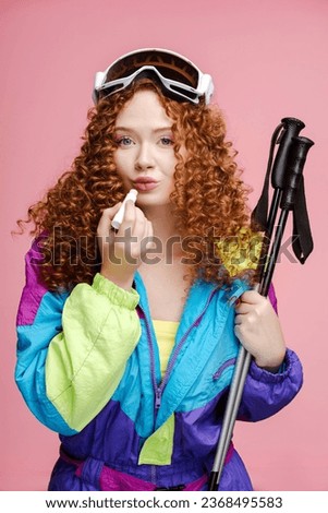 Portrait of fashion curly woman wearing ski goggles holding lipstick applying lips looking in mirror isolated on pink background. Beauty concept