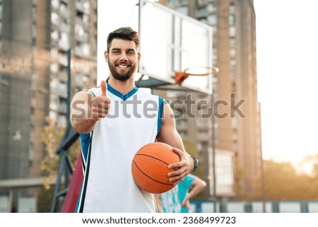 Young handsome male basketball player holding ball and thumbs up while smiling and looking at camera outdoors. Urban background with basketball court.
