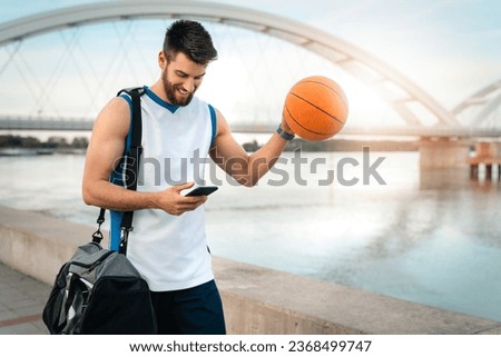 Handsome basketball player feeling happy and satisfied while looking at mobile phone walking by the river with sports equipment. Professional basketball player athlete using smart phone.