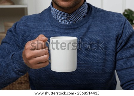 Man holding white mug indoors, closeup. Mockup for design Royalty-Free Stock Photo #2368514415