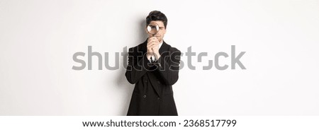 Image of serious businessman in black trendy suit, looking through magnifying glass, looking for employees, standing against white background.