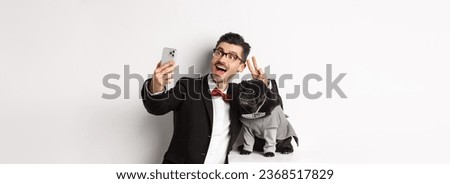 Cheerful dog owner in suit celebrating New Year with dog, taking selfie on smartphone near cute black pug in costume, white background.
