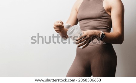Athletic woman in sportswear with measuring spoon in her hand puts a portion of whey protein powder into a shaker, making protein drink, cropped image. Royalty-Free Stock Photo #2368536653